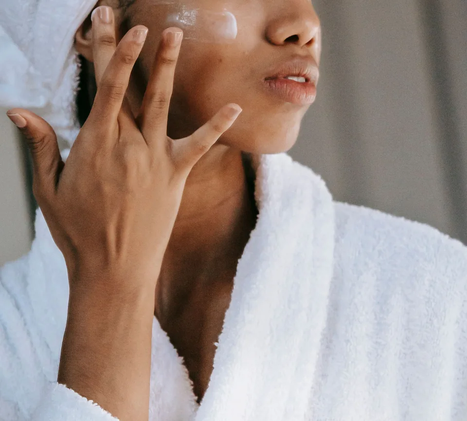 Fingers gently spreading cream on a cheek above a white robe.