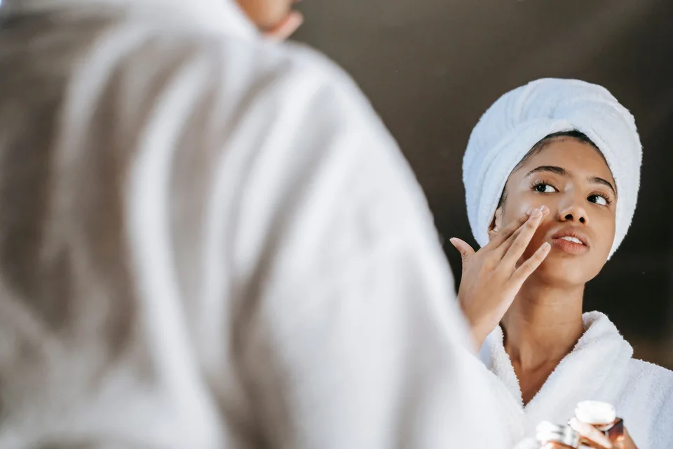 A woman in a white robe applies cream to her face while looking in a mirror.